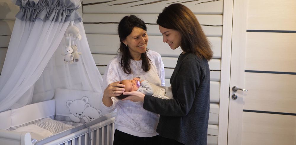 Mother holding newborn baby girl in her arms stroking her head talking and laughing to a friend in the baby's beautiful nursery